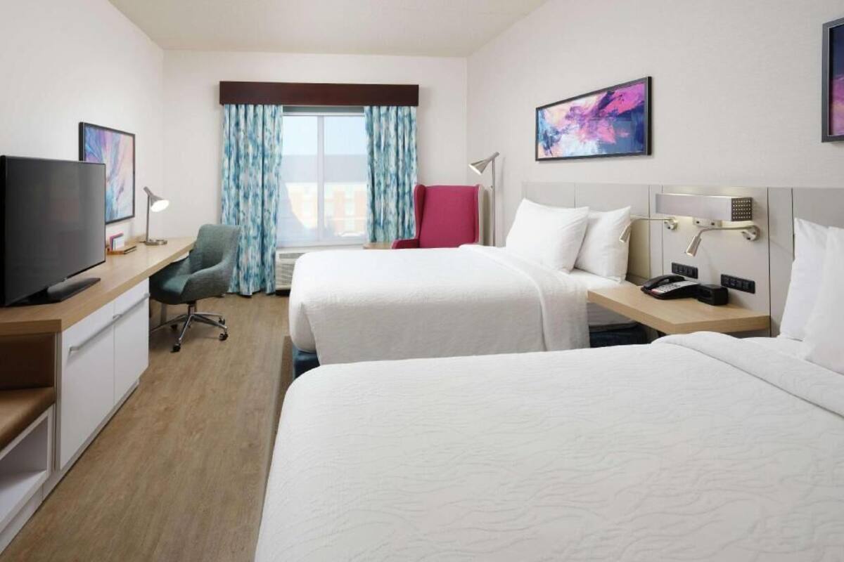 A hotel room with two beds, a desk with a TV, a chair, a desk lamp, and a window with curtains.