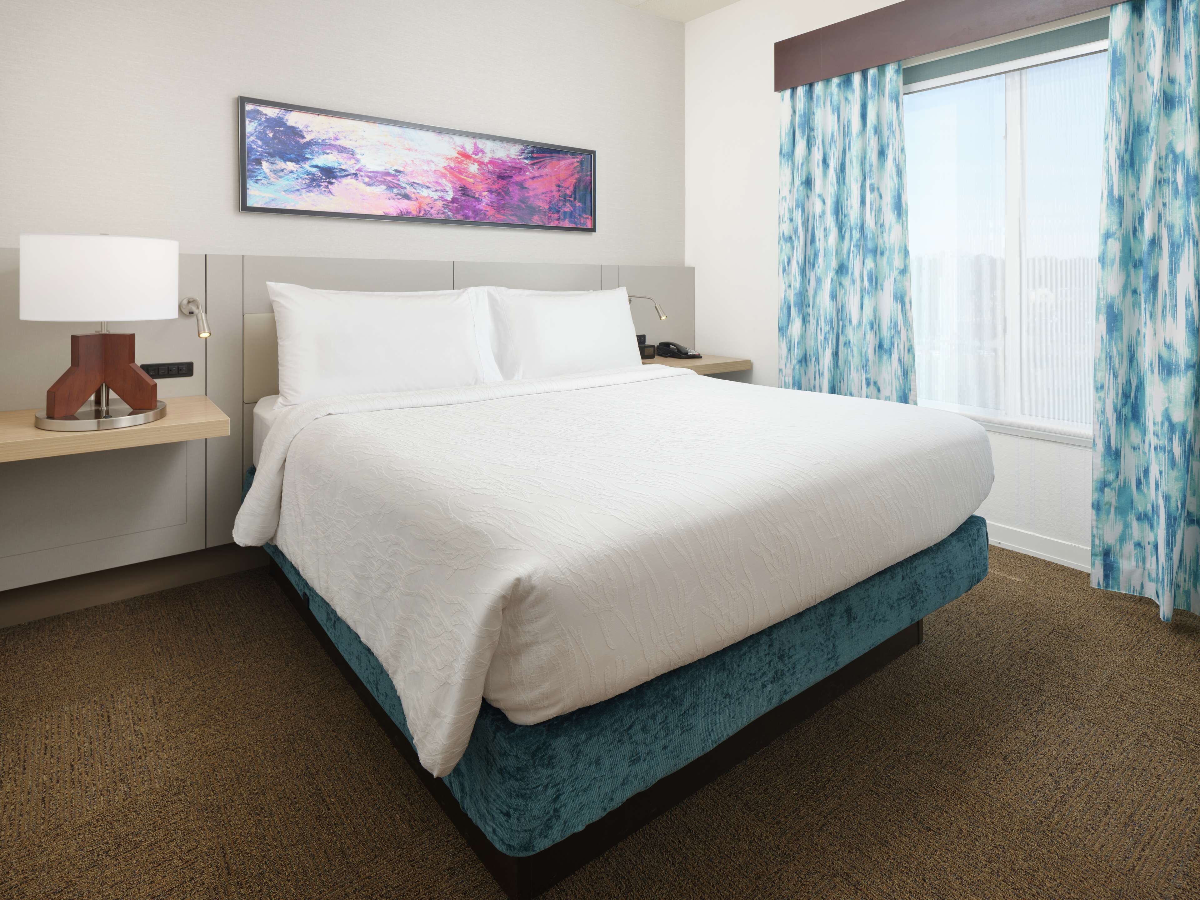 A hotel room with a large bed, a bedside table with a lamp, a painting on the wall, and a window with curtains.