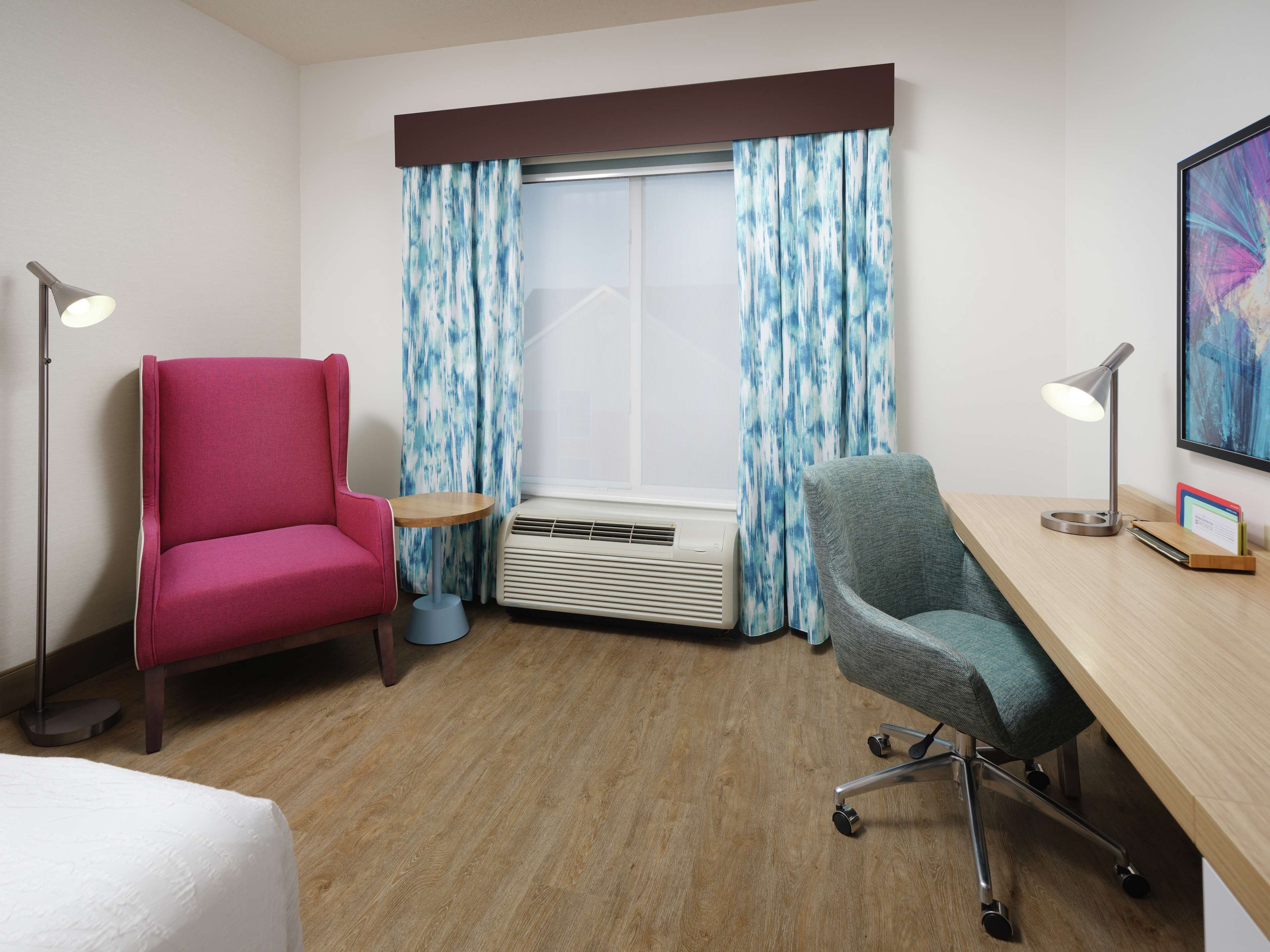 A hotel room with a wooden floor, a desk with a chair, a red armchair, a standing lamp, and a window with blue curtains.