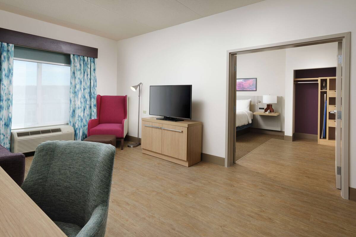 A hotel room with a wooden desk, a red armchair, a television on a wooden cabinet, a floor lamp, and a view of another room through an open door.