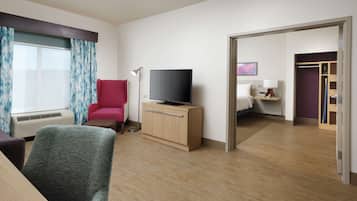 A hotel room with a wooden desk, a red armchair, a television on a wooden cabinet, a floor lamp, and a view of another room through an open door.