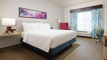 Una camera d'albergo con un letto grande, una scrivania con lampada, una sedia, una finestra con tende e un quadro appeso al muro.