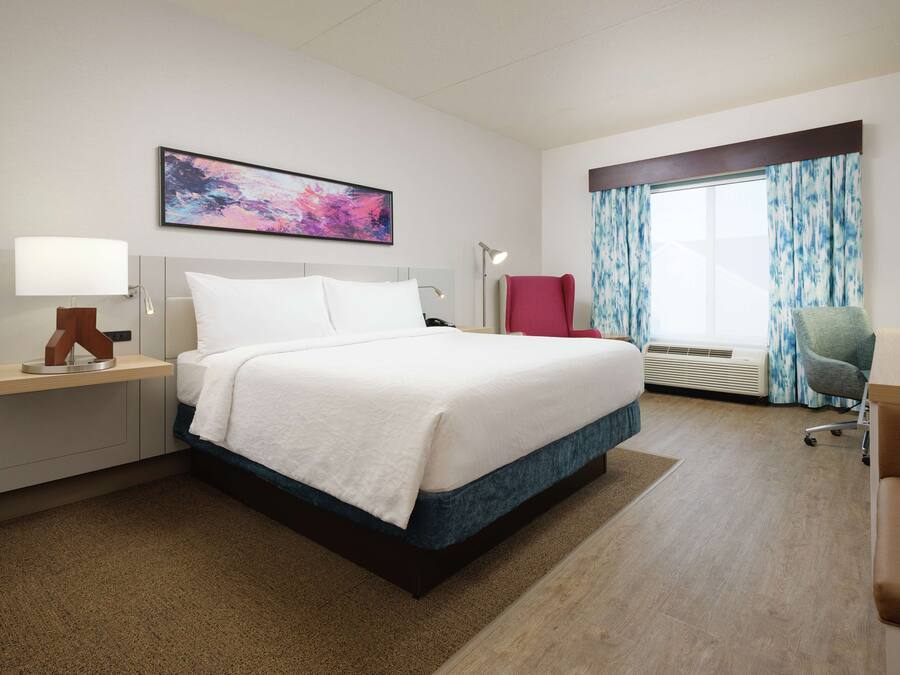 Una camera d'albergo con un letto grande, una scrivania con lampada, una sedia, una finestra con tende e un quadro appeso al muro.