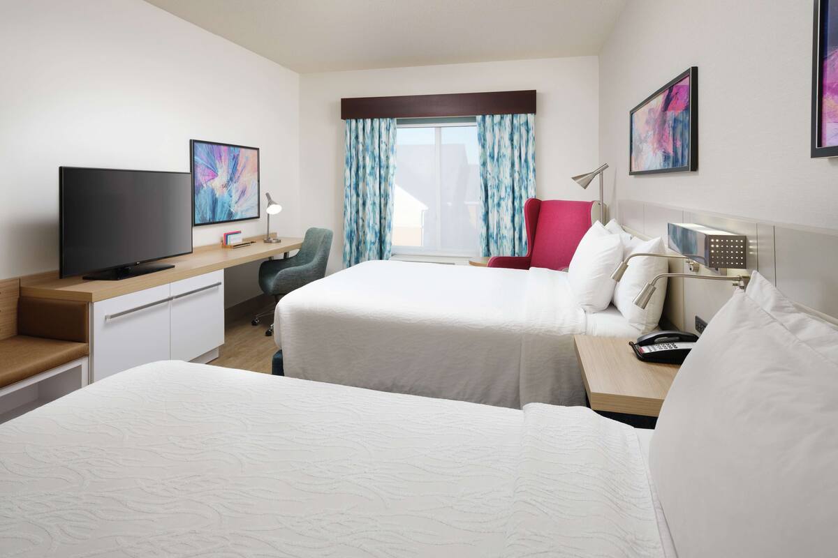 A hotel room with two beds, a desk with a TV, a chair, a desk lamp, and artwork on the walls.