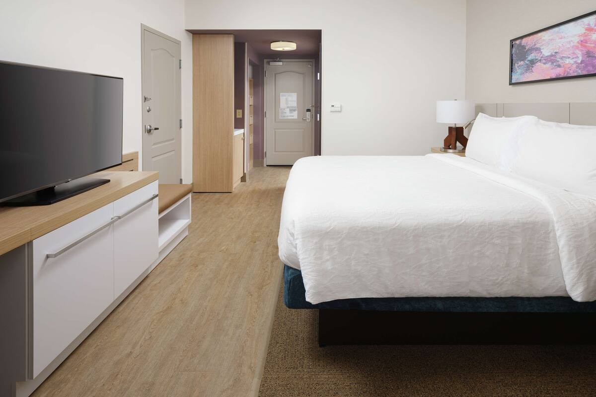 A hotel room with a bed, a TV, a desk, and a painting on the wall.