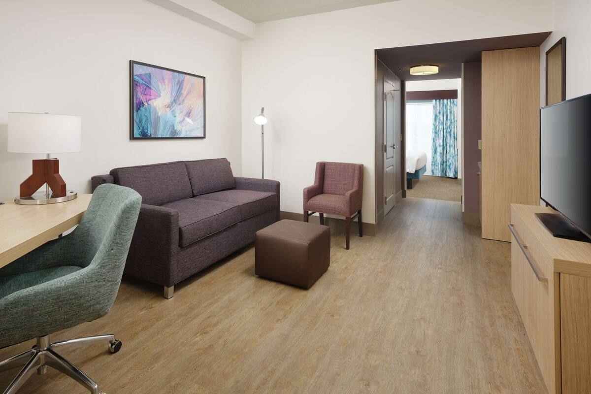 A modern hotel room with a wooden floor, a sofa, a chair, a desk, a lamp, a TV, and a painting on the wall.