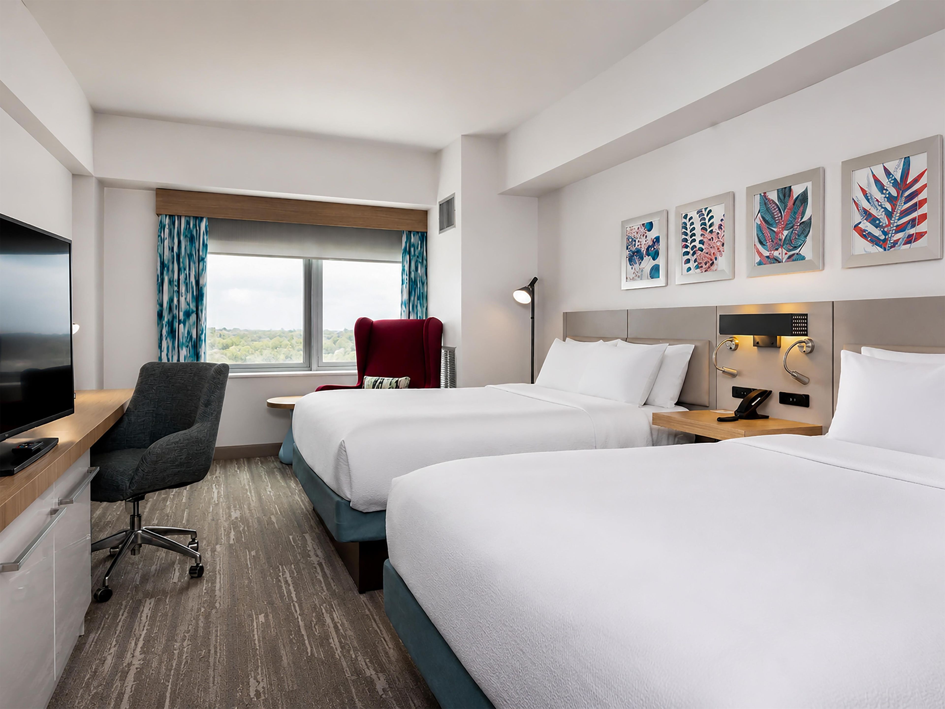 Ett hotellrum med två sängar, ett skrivbord med en tv, en stol, ett fönster med gardiner och konstverk på väggen.