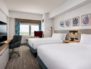 Habitación de hotel con dos camas, un escritorio con televisor, una silla, un ventanal con cortinas y obras de arte en la pared.