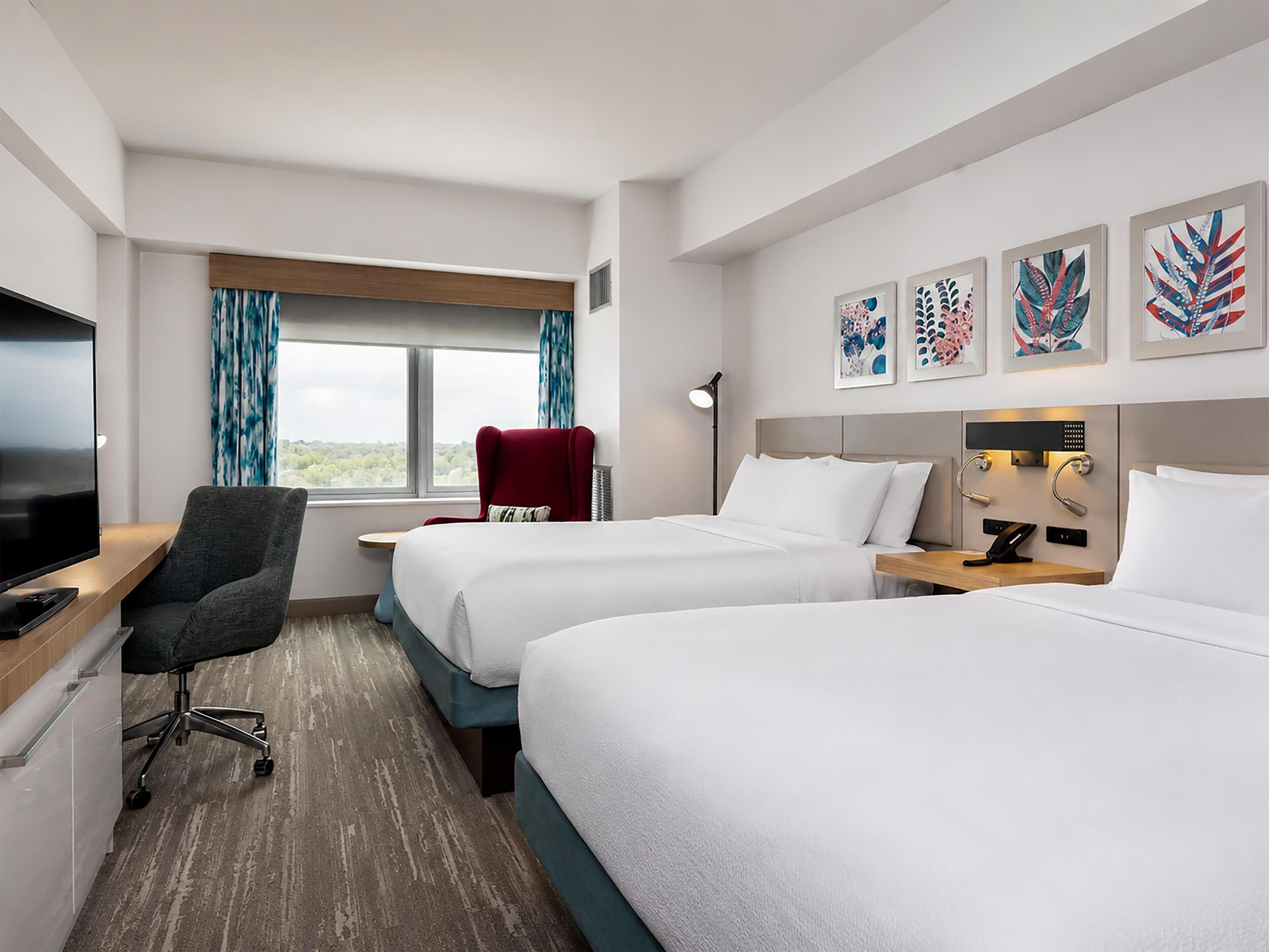 Habitación de hotel con dos camas, un escritorio con televisor, una silla, un ventanal con cortinas y obras de arte en la pared.
