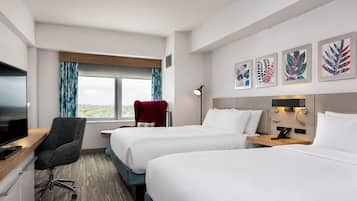Habitación de hotel con dos camas, un escritorio con televisor, una silla, un ventanal con cortinas y obras de arte en la pared.