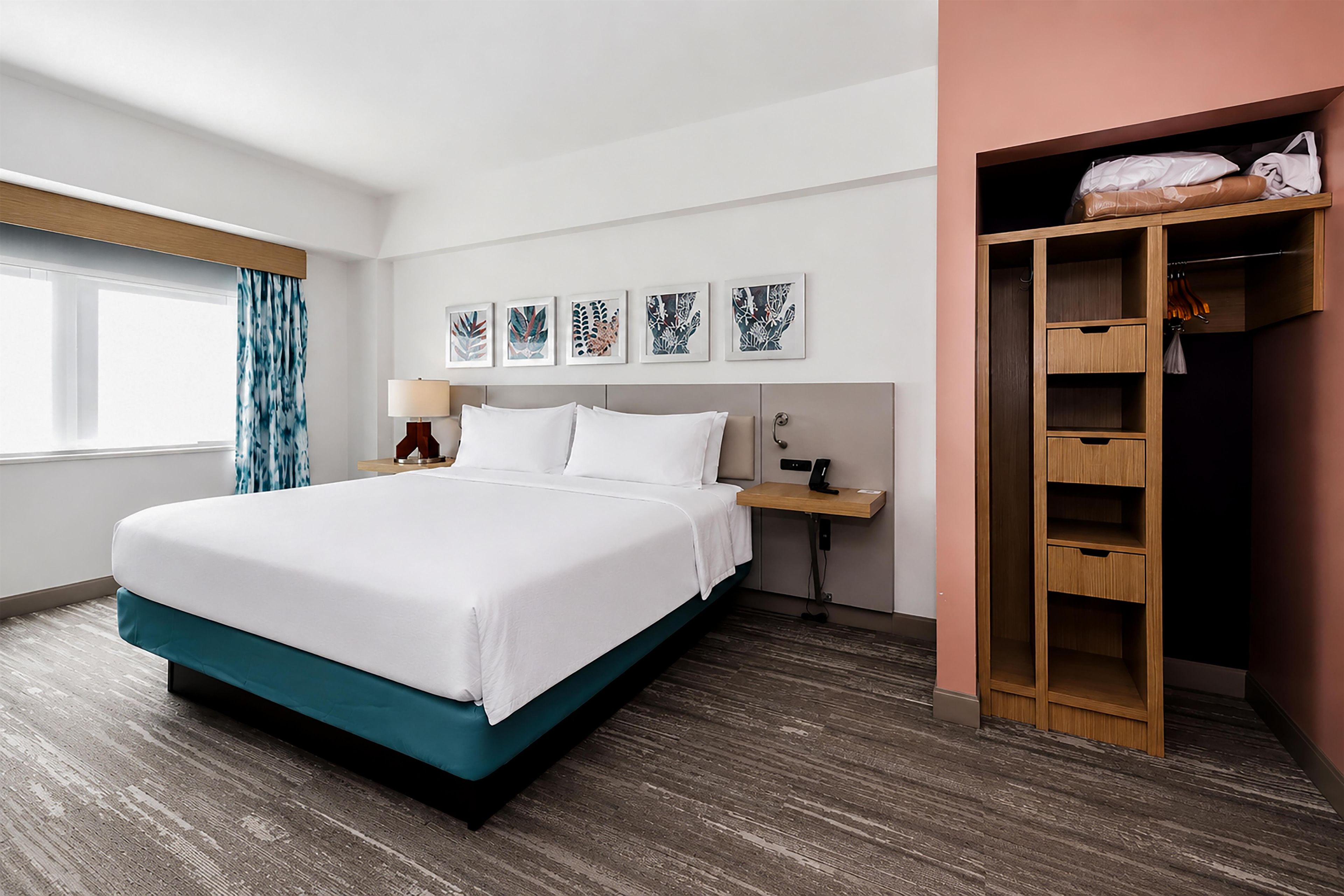 A hotel room with a bed, a nightstand, a lamp, a wall-mounted phone, a window with curtains, and a wooden wardrobe.