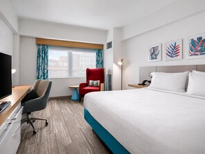 Habitación de hotel con cama, escritorio con computadora, silla, ventana con cortinas, mesita de noche con lámpara y una obra de arte en la pared.