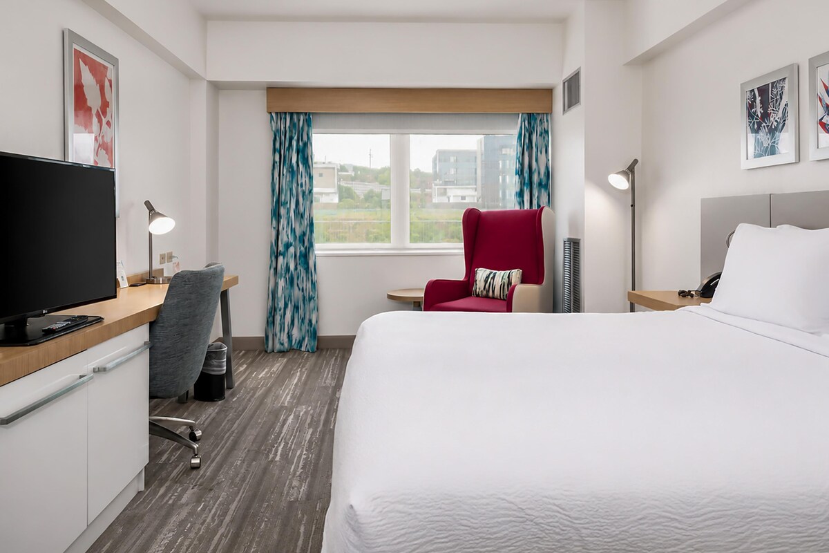 Une chambre d’hôtel avec un lit, un bureau avec un ordinateur, une chaise, une télévision et une fenêtre avec des rideaux.