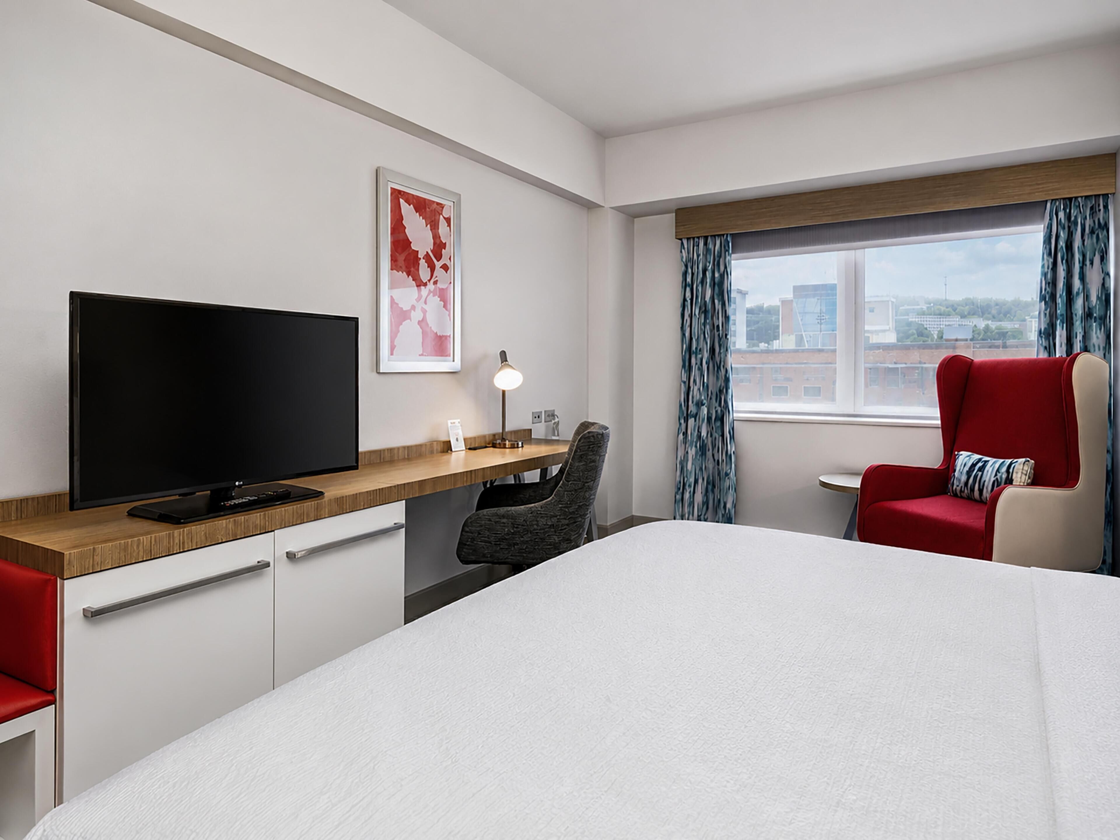 A hotel room with a bed, a desk with a TV, a chair, a window with curtains, and a city view.