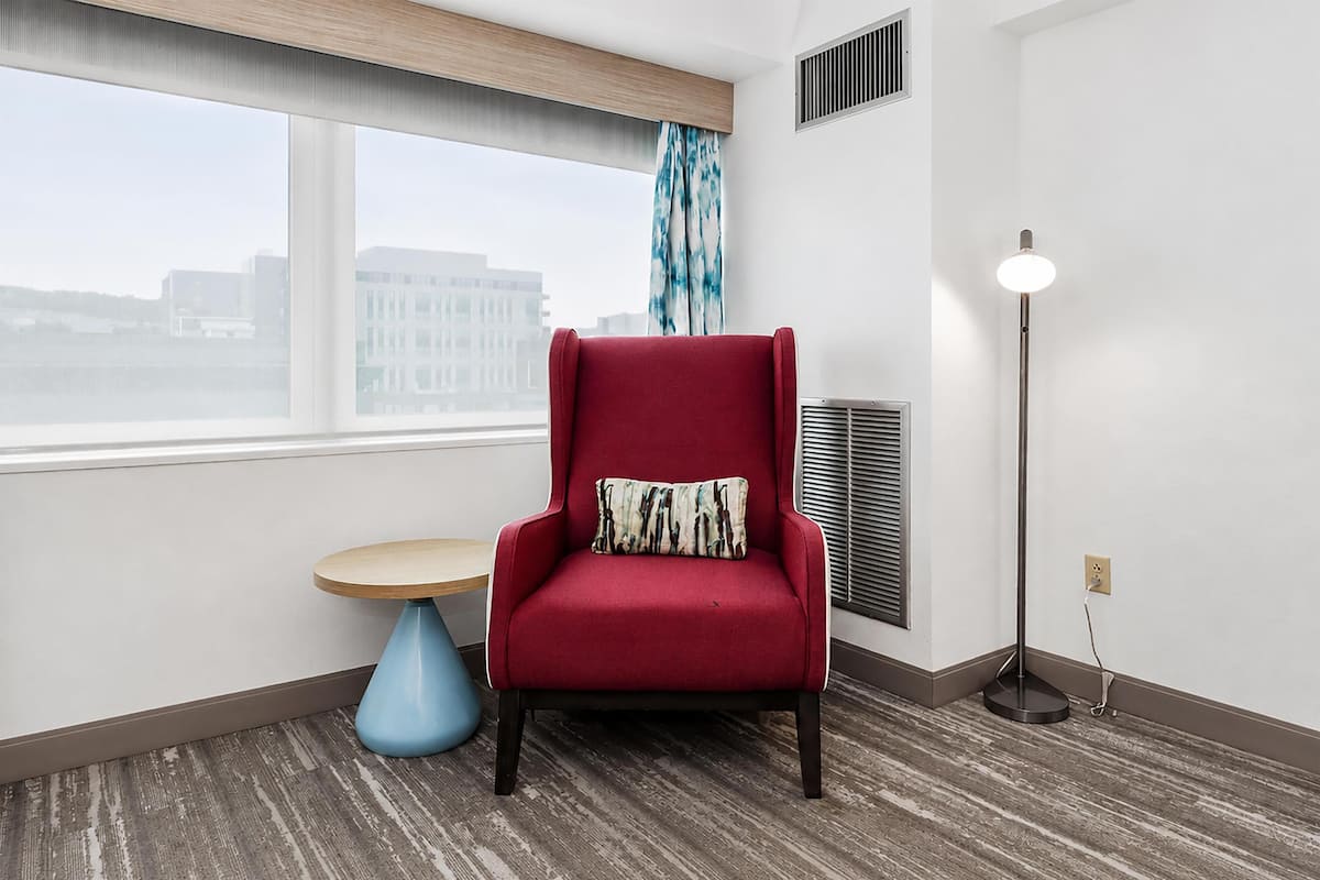 A red armchair with a patterned pillow, a round wooden table, a floor lamp, and a window with a view of buildings.