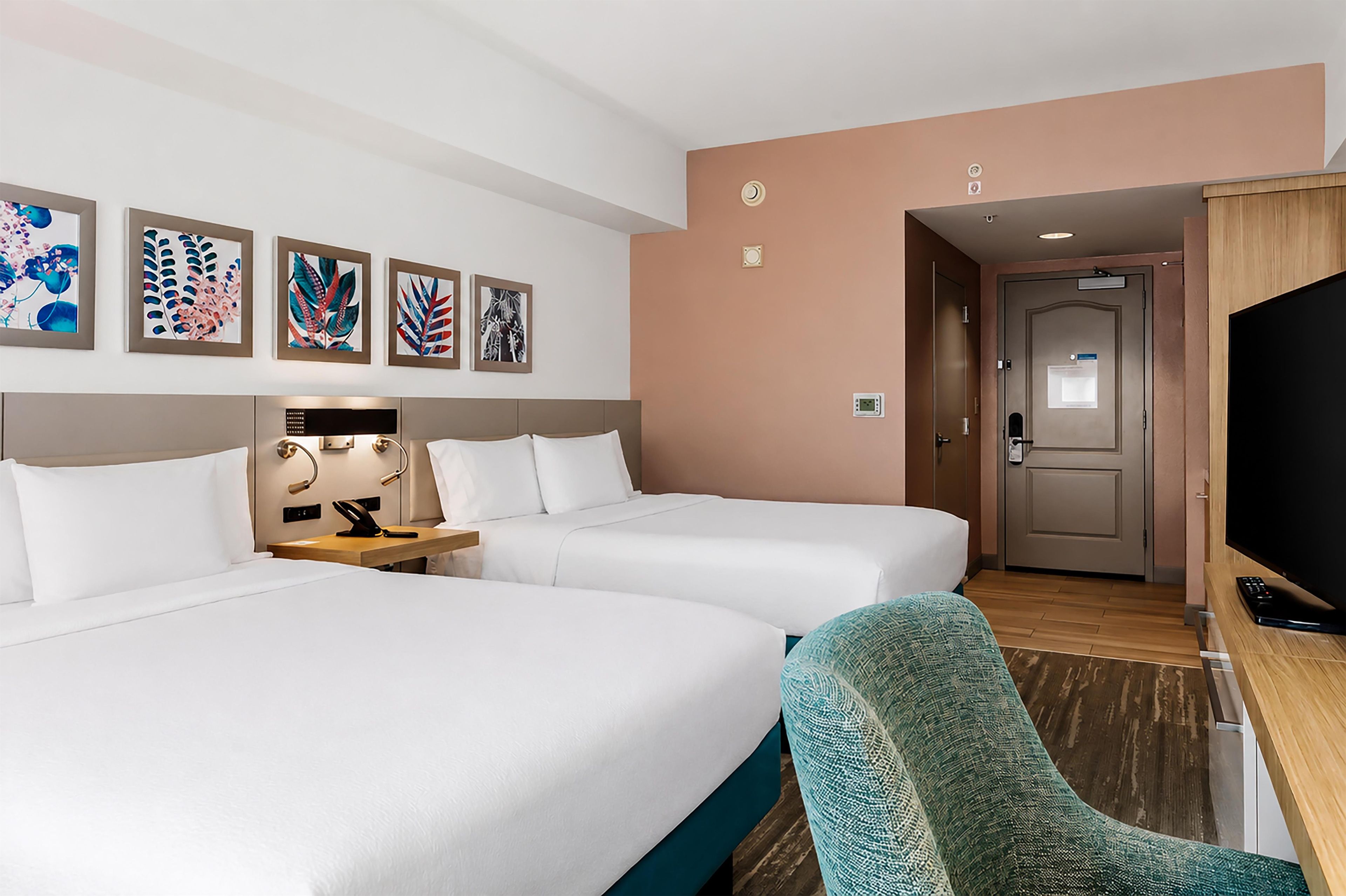 A hotel room with two beds, a desk with a lamp, a television, and framed artwork on the wall.