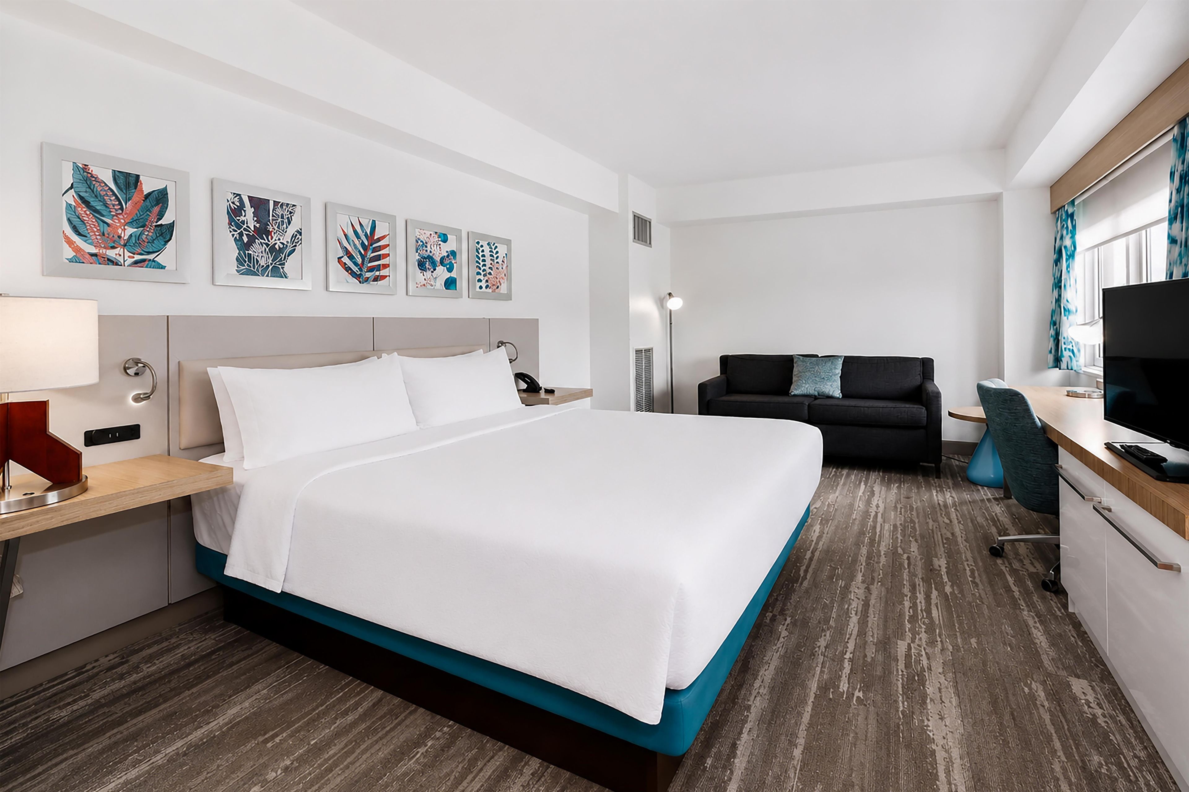 A hotel room with a bed, a desk, a chair, a TV, and artwork on the wall.