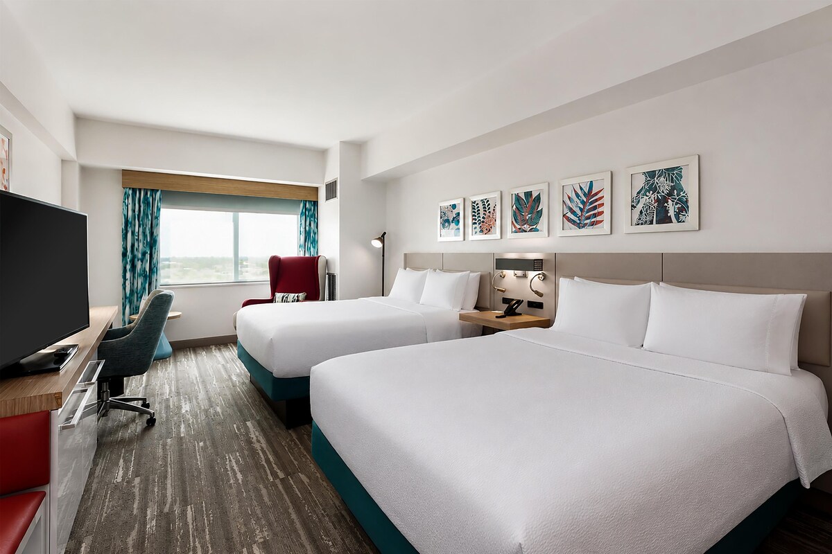 Une chambre d’hôtel avec deux lits, un bureau avec une télévision, une chaise, une fenêtre avec des rideaux et des œuvres d’art au mur.