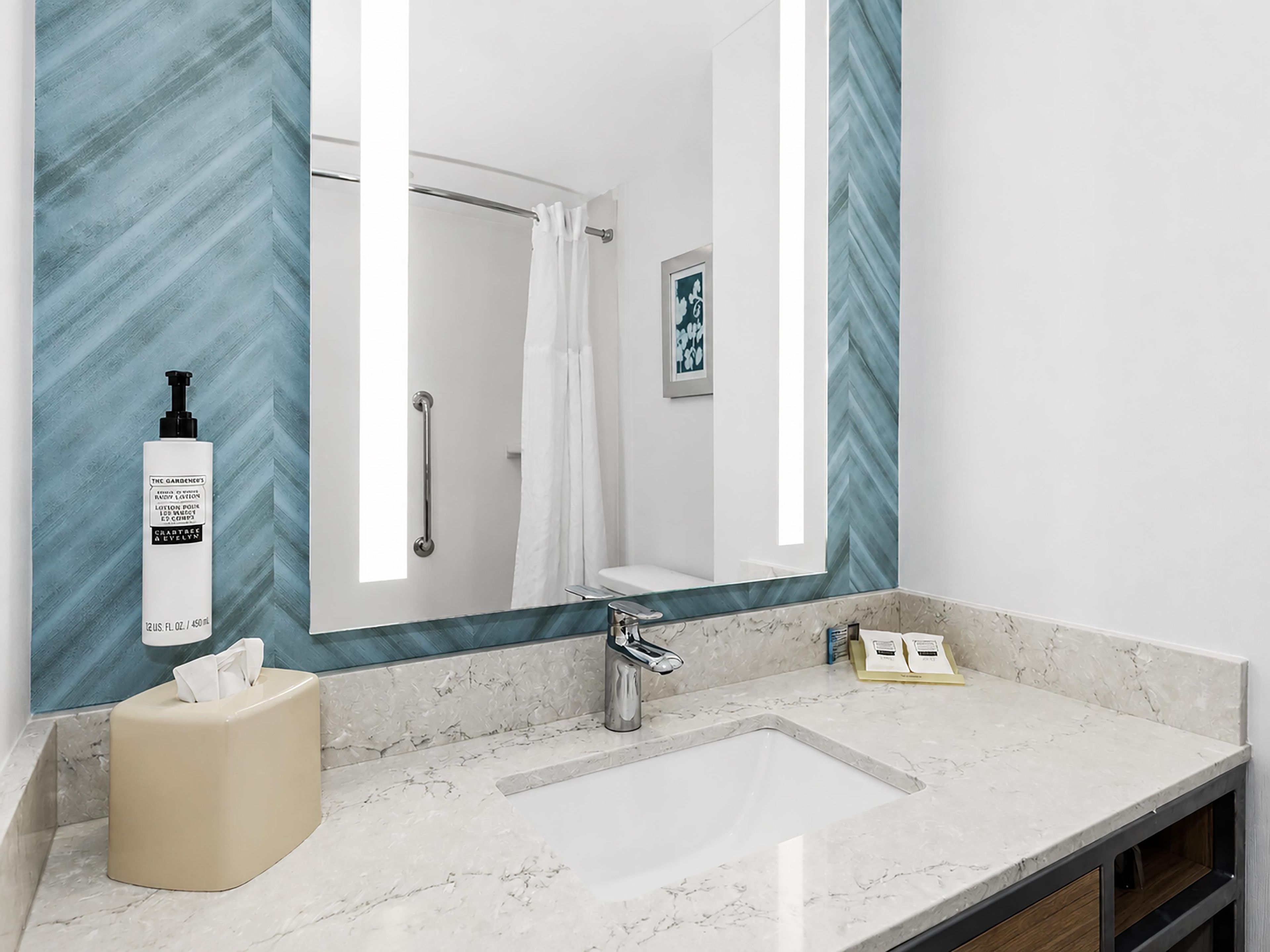 A bathroom with a large mirror, a sink, a towel dispenser, and a shower curtain.