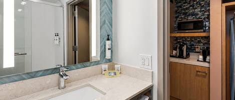 A bathroom with a large mirror, a sink, and a shower. There is a towel rack and a bottle of shampoo on the counter.