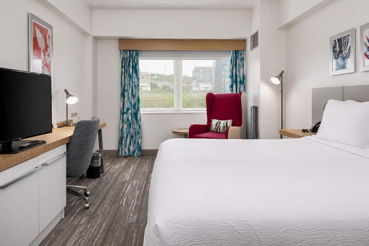 A hotel room with a bed, a desk with a computer, a chair, a TV, and a window with curtains.