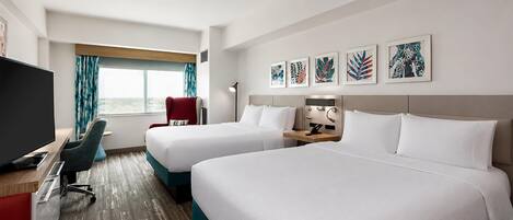 A hotel room with two beds, a desk with a TV, a chair, a window with curtains, and artwork on the wall.
