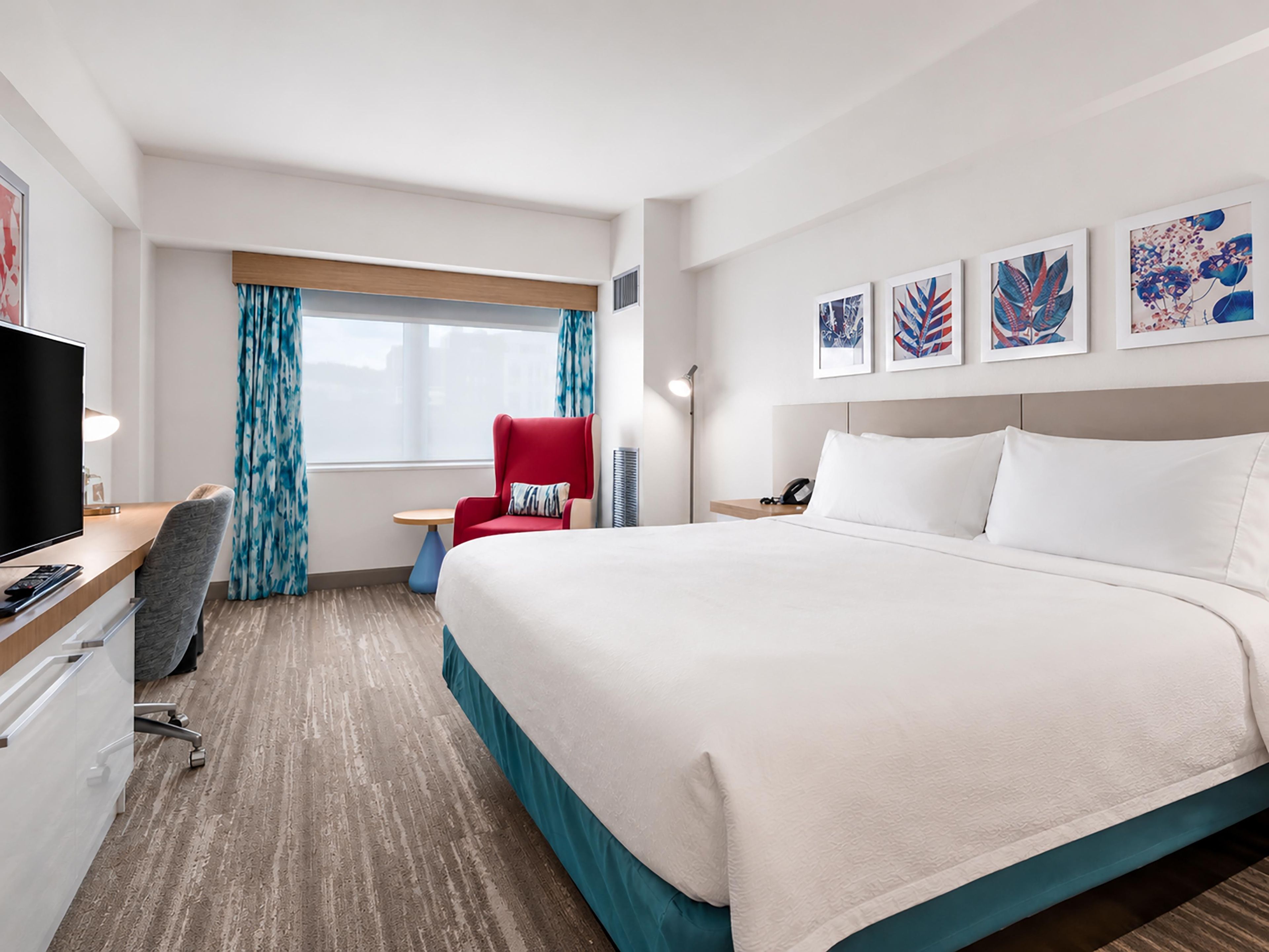 A hotel room with a bed, a desk with a TV, a chair, a small table, and artwork on the wall.