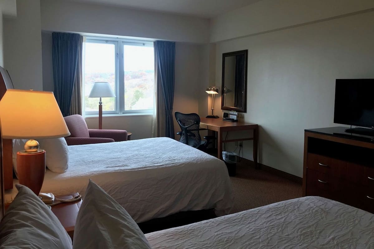 A hotel room with two beds, a desk with a lamp, a television, and a window with curtains.