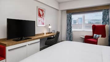 A hotel room with a bed, a desk with a TV, a chair, a window with curtains, and a city view.