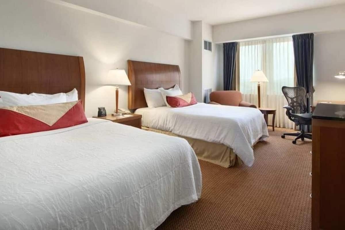 A hotel room with two beds, a desk, a chair, and a lamp.