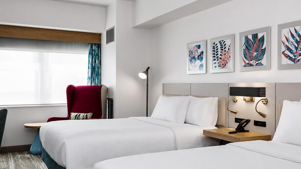 A hotel room with two beds, a red armchair, a floor lamp, and artwork on the wall. - Hilton Garden Inn Ithaca (Ithaca)