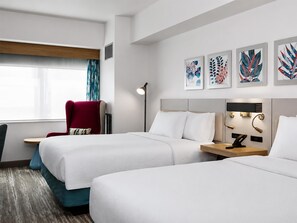 Quarto de hotel com duas camas, uma poltrona vermelha, uma luminária de chão e obras de arte na parede.