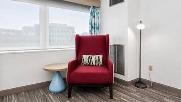 A red armchair with a patterned pillow, a round wooden table, a floor lamp, and a window with a view of buildings.
