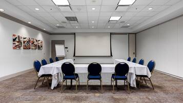 A conference room with a long table, blue chairs, a projector screen, and artwork on the wall.
