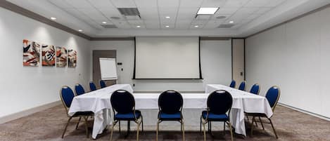 A conference room with a long table, blue chairs, a projector screen, and artwork on the wall.