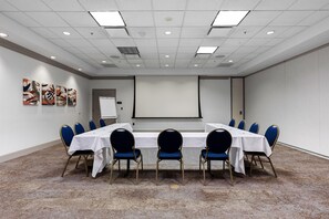 A conference room with a long table, blue chairs, a projector screen, and artwork on the wall. - Hilton Garden Inn Ithaca (Ithaca)