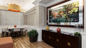 A modern living room with a dining area, a large window with artwork, a dark wood cabinet, and a potted plant. - Hilton Garden Inn Ithaca (Ithaca)