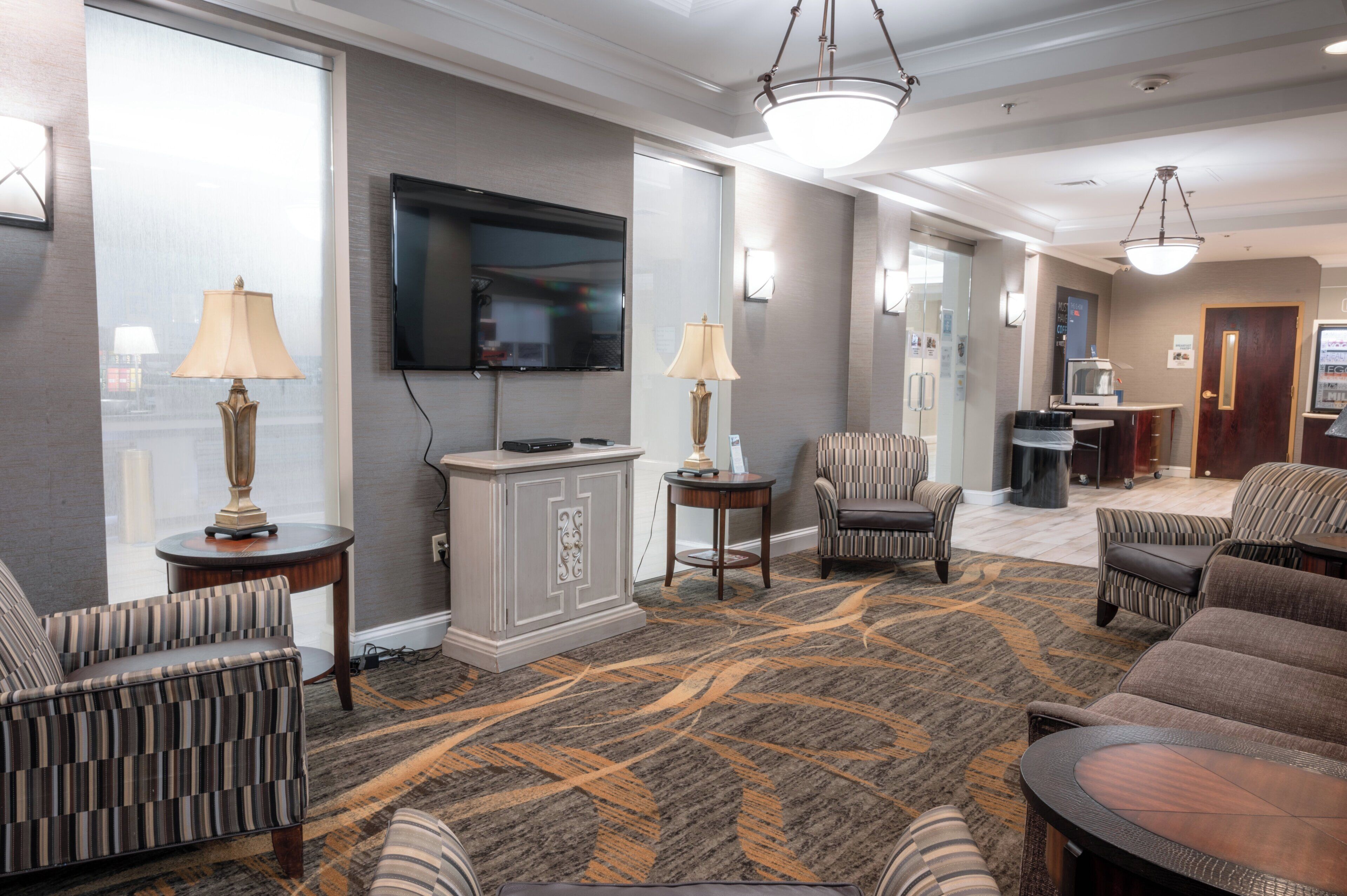 A hotel lobby with a large flat-screen TV, multiple seating areas, a patterned carpet, and a kitchenette.