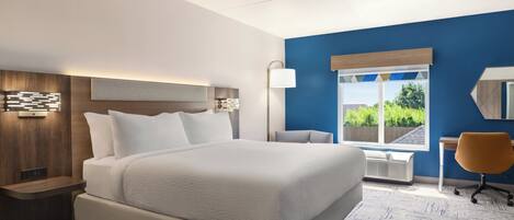 A modern hotel room with a large bed, a desk, a chair, and a view of greenery outside.