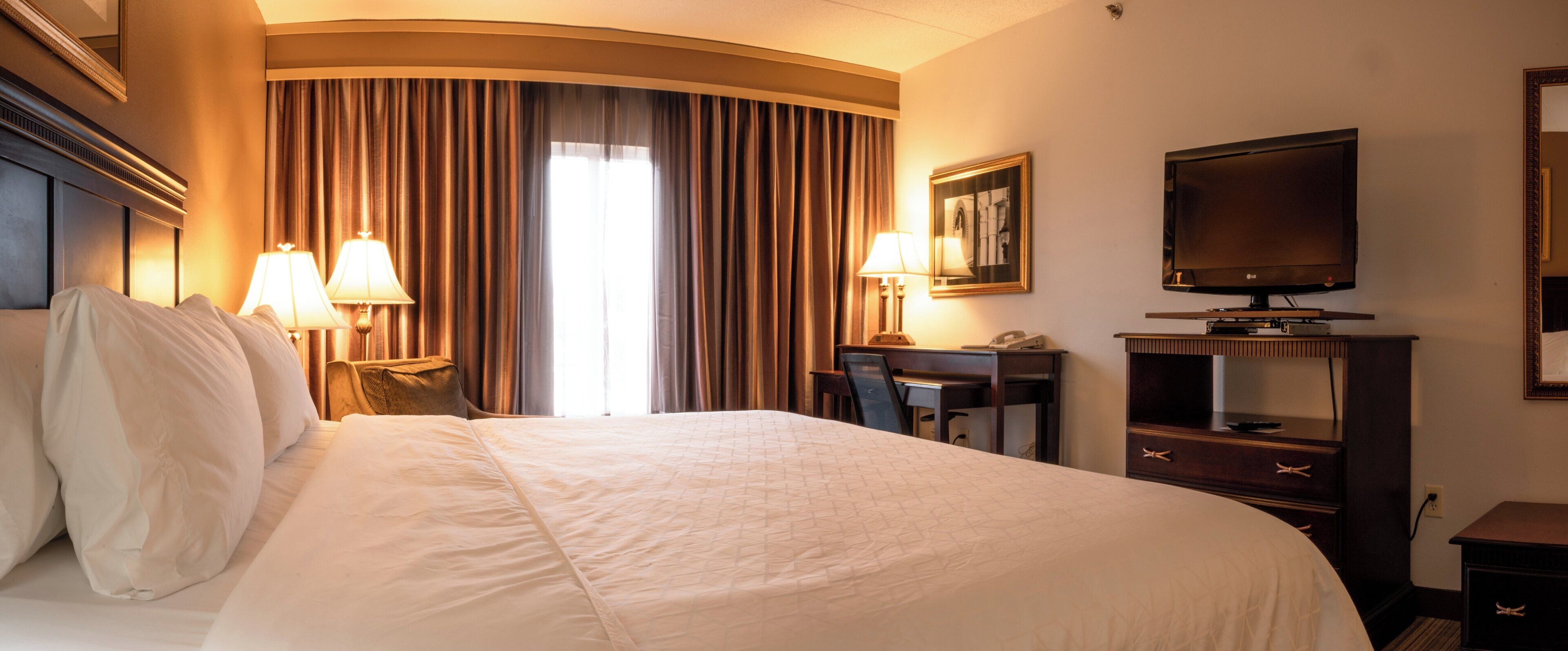 A hotel room with a large bed, two bedside lamps, a desk with a chair, a television, and a framed picture on the wall.