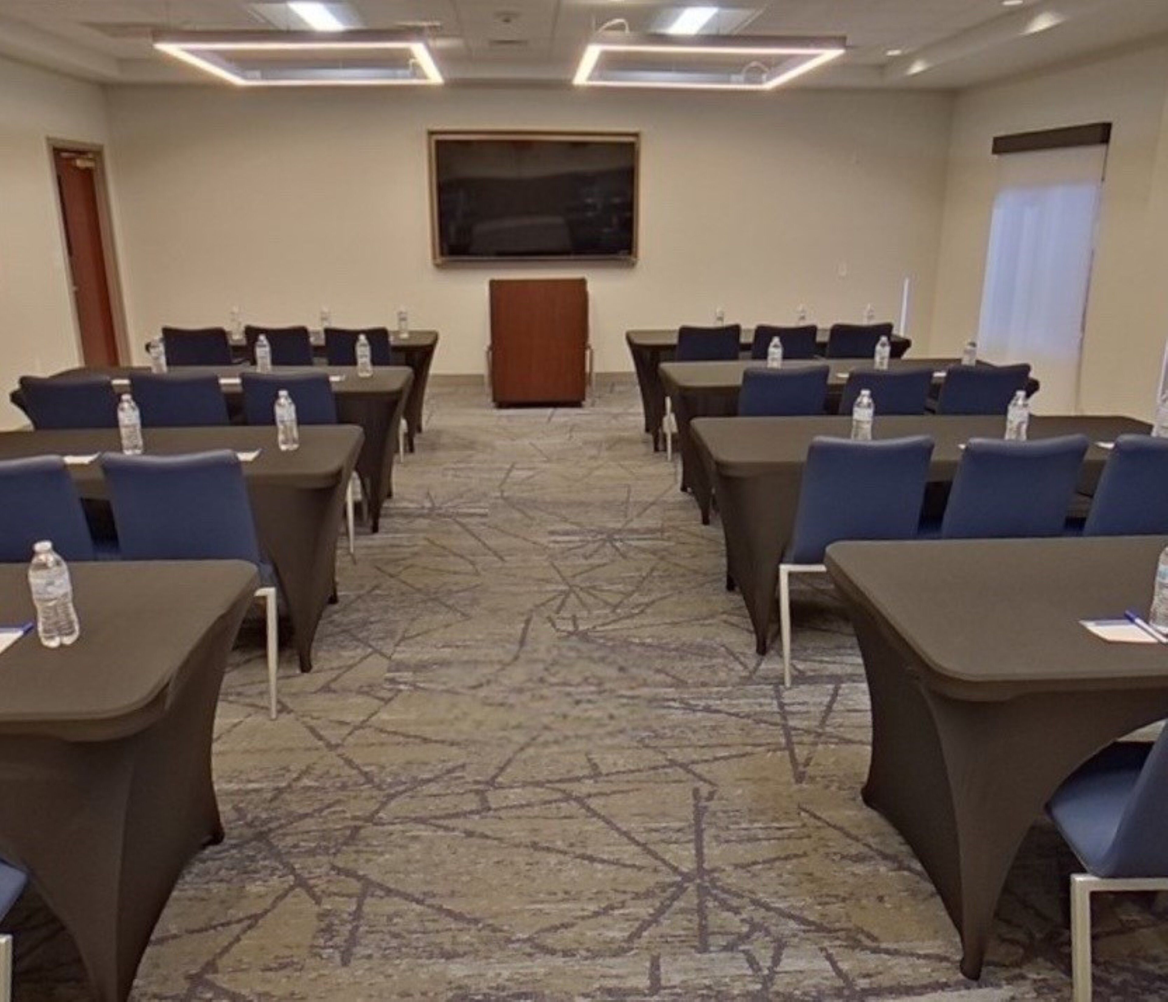 Une salle de conférence équipée de tables et de chaises, dotée d’un sol recouvert de moquette et d’un grand écran situé au fond de la pièce.