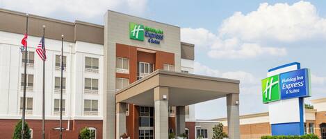 Edificio de Holiday Inn Express & Suites con un acceso cubierto, banderas estadounidenses y un área ajardinada con plantas.