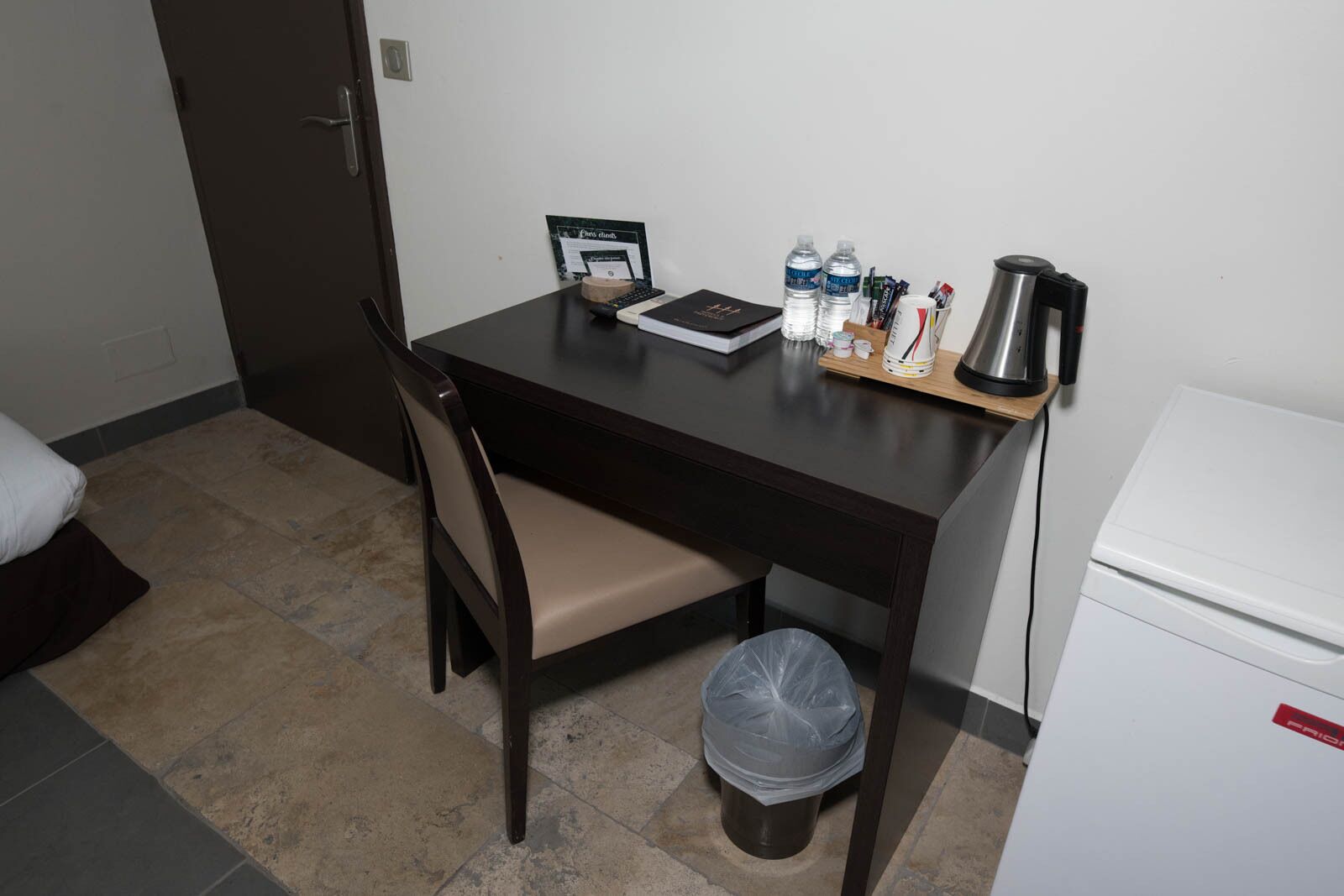 A small, dark wooden desk with a chair, a laptop, water bottles, a coffee maker, and a trash can.