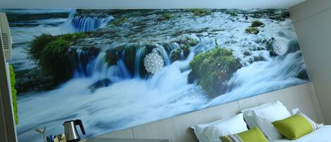 Une chambre avec une grande fresque murale représentant une cascade, un lit avec des oreillers blancs et verts, et une table de chevet sur laquelle se trouvent une bouilloire et des tasses.