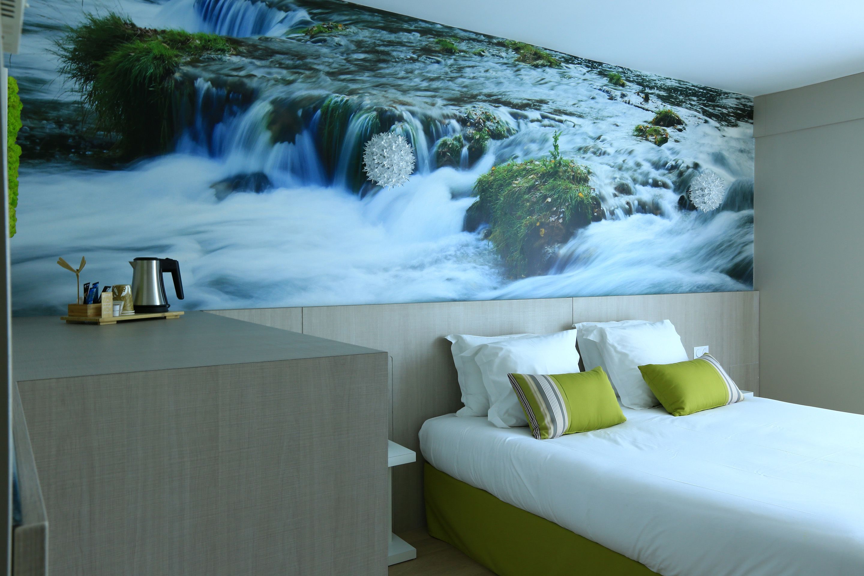 Ein Hotelzimmer mit einer großen Wandmalerei eines Wasserfalls, einem Bett mit weißer und grüner Bettwäsche sowie einem Nachttisch mit Wasserkocher und Tassen.