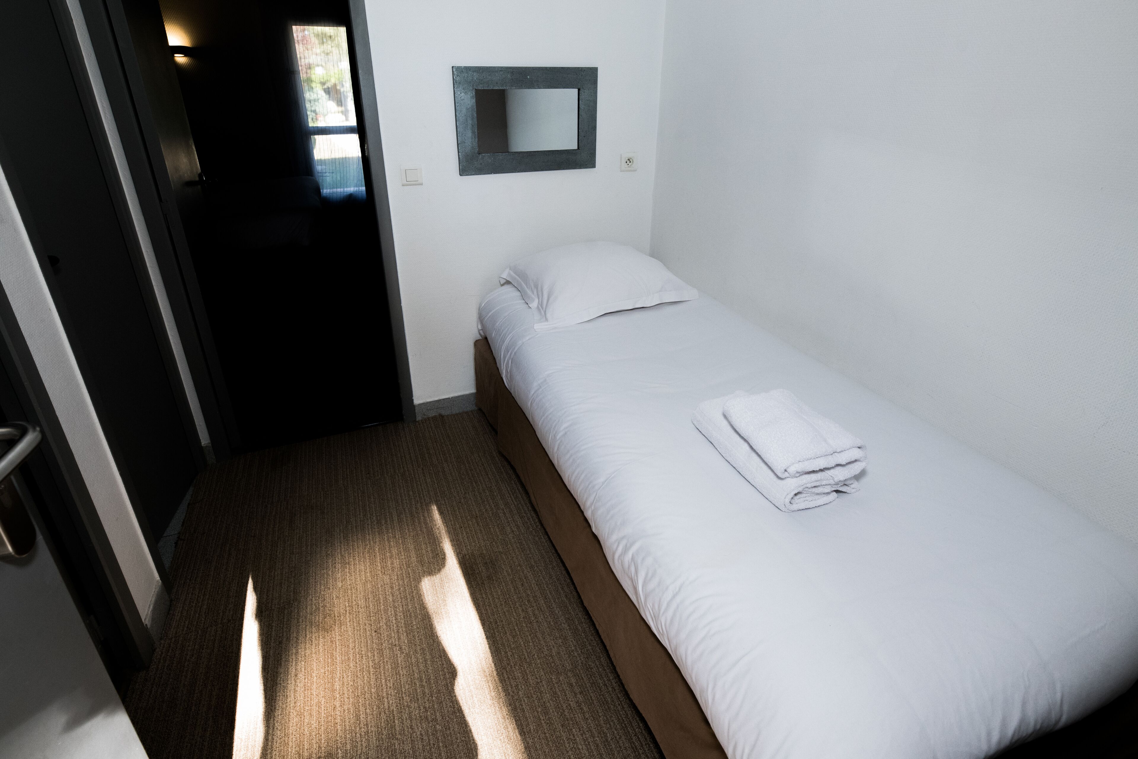 Un petit lit bien fait, avec du linge de lit blanc et une serviette pliée, un seul miroir fixé au mur et un sol recouvert d’une moquette.