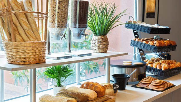 Mostrador de panadería con diferentes panes y pasteles, una máquina de café y una planta en maceta.