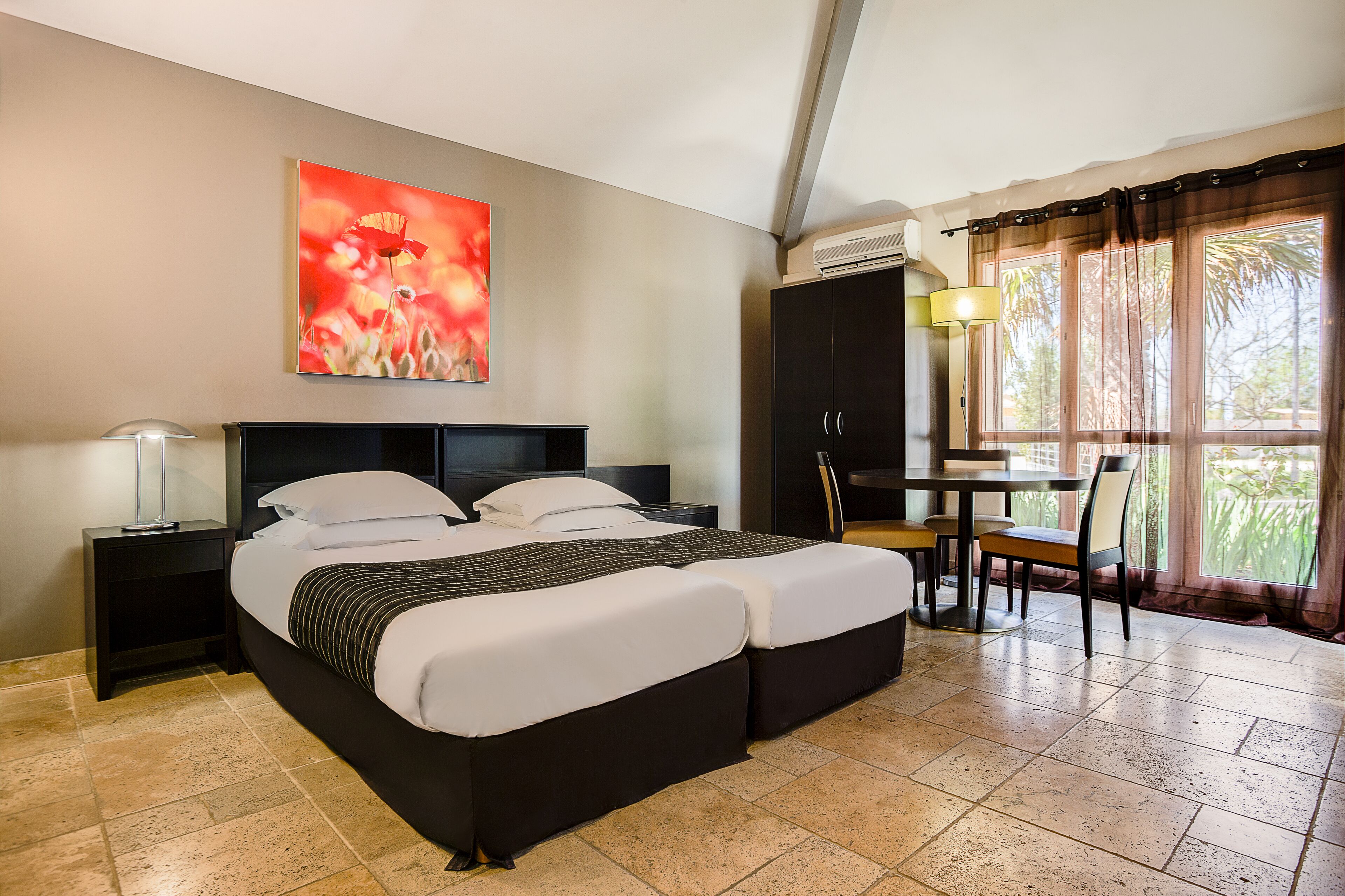 A hotel room with a bed, a desk, and a chair. There is a painting on the wall and a window with curtains.