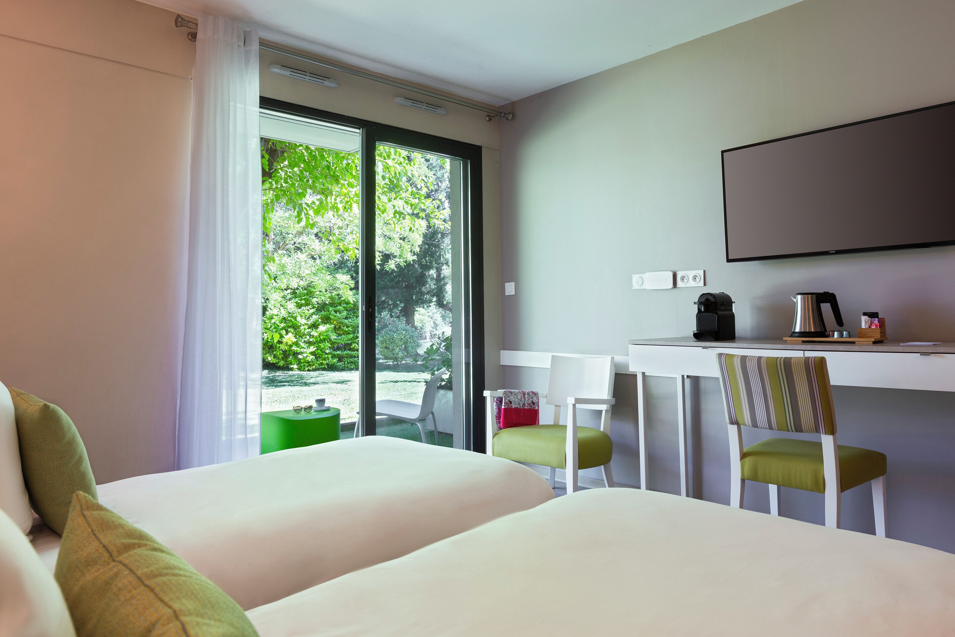 A hotel room with two beds, a desk, and a chair. There is a large window with a view of greenery outside.