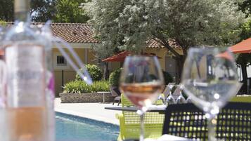 Une table placée au bord de la piscine, avec un verre de vin et une bouteille, entourée de sièges d’extérieur et d’un bâtiment dont le toit est recouvert de tuiles.